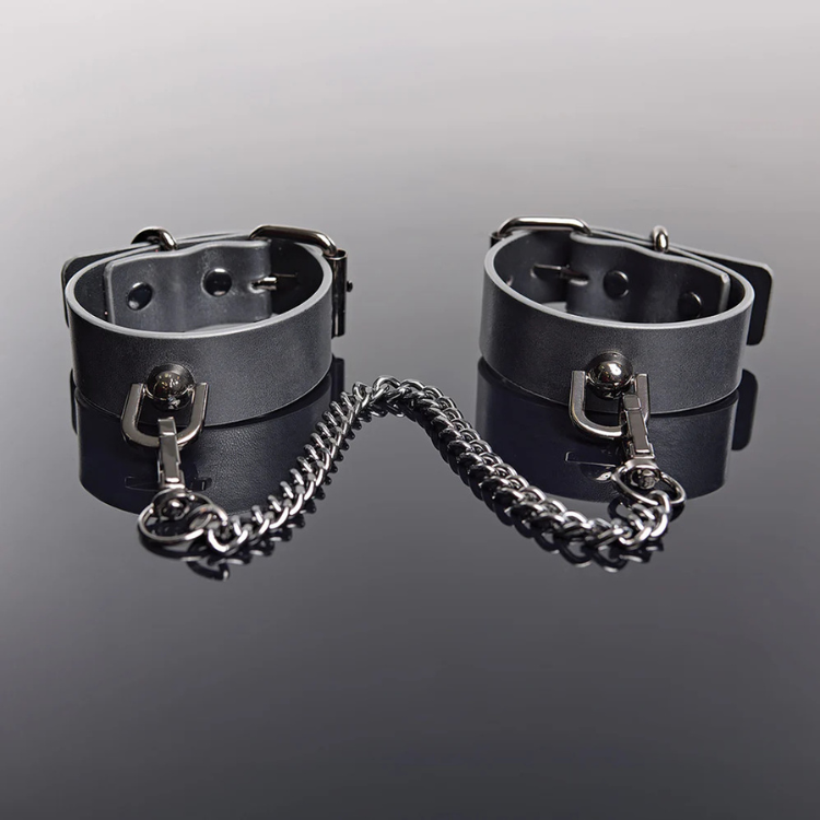 Grey faux leather pleasure cuffs with silver chain on reflective surface, House of Vices wellness essentials
