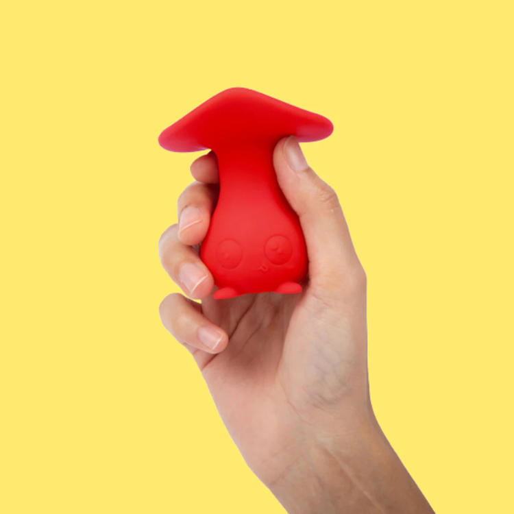 Hand holding a cute red mushroom-shaped silicone wellness toy on a yellow background