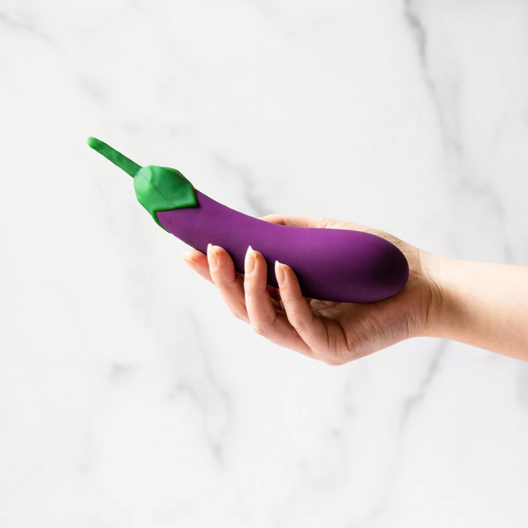 Hand holding a purple eggplant-shaped wellness toy against a marble background, House of Vices